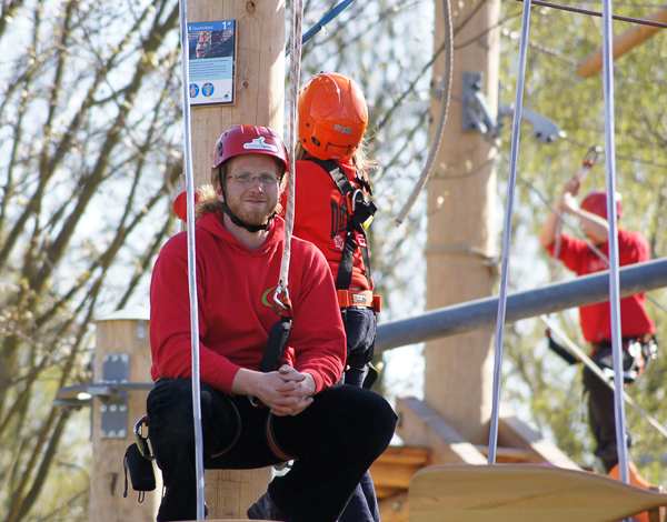 Werde Kletterpark Manager (m/w/d) im HanseRock Junger Mitarbeiter in Kletterausrüstung schaut von einem Kletterelement freundlich in die Kamera